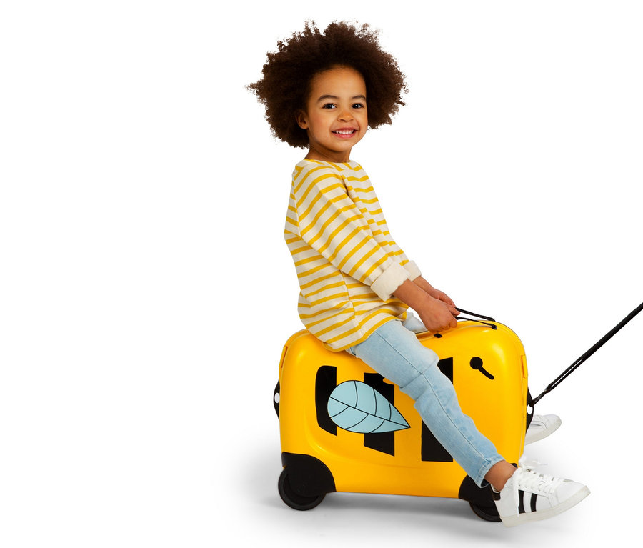 Ein lächelndes Kind mit lockigem Haar sitzt auf einem gelben American Tourister Funrider Aufsitzkoffer im Bienen-Design.