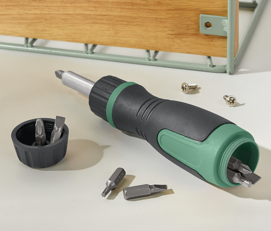 Ein schwarz-grüner magnetischer Ratschenschraubendreher liegt neben einem Deckel mit Bits und zwei Schrauben.