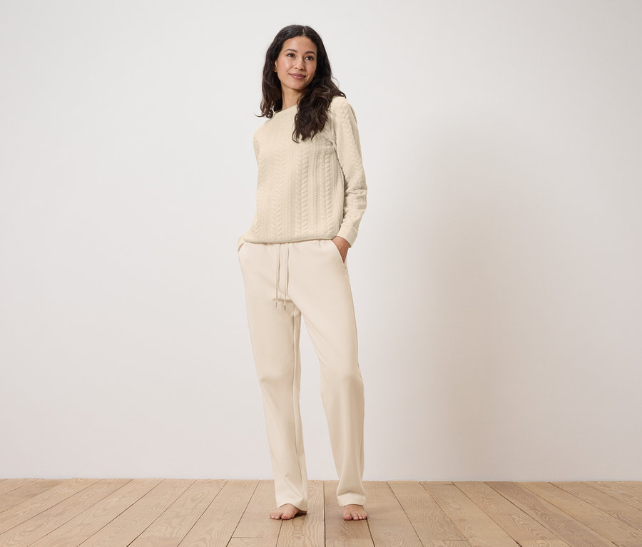 Eine Frau steht mit den Händen in den Taschen einer cremefarbenen Jogginghose mit einem cremefarbenen Zopfmusterpullover.