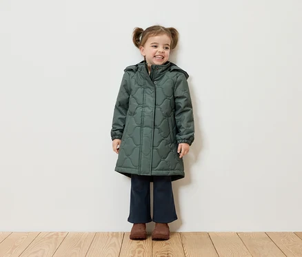 Ein lächelndes Mädchen mit Zöpfen steht vor einer weißen Wand und trägt einen grünen Mantel, dunkelblaue Hosen und braune Schuhe.