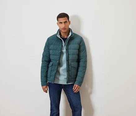 Mann posiert vor einem weißen Hintergrund in einer blauen Jacke, blauem Pullover und Jeans.