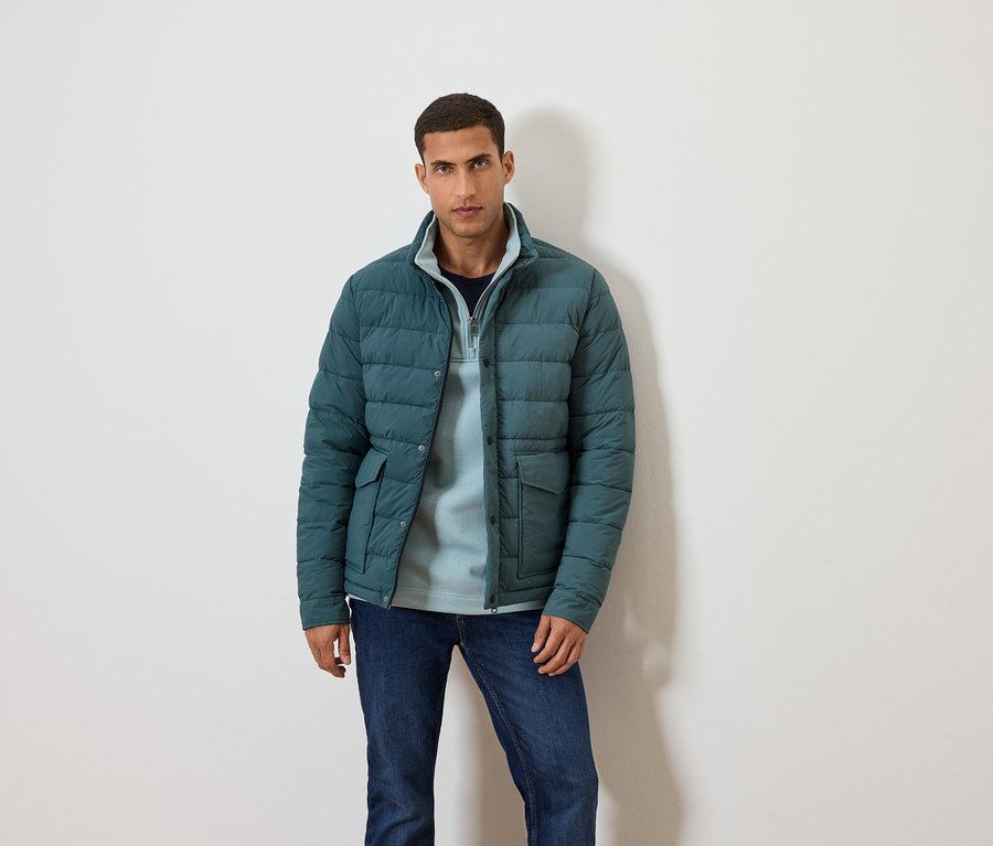 Mann posiert vor einem weißen Hintergrund in einer blauen Jacke, blauem Pullover und Jeans.