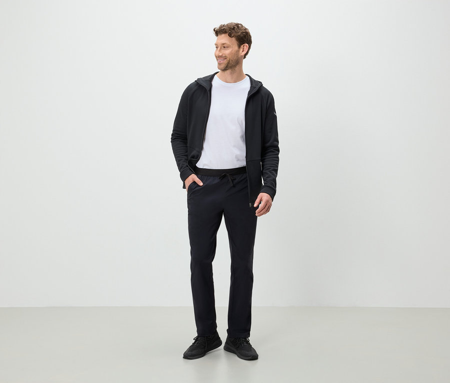 Ein Mann steht und schaut zur Seite, er trägt eine schwarze Hose, ein weißes T-Shirt und eine schwarze Kapuzenjacke.