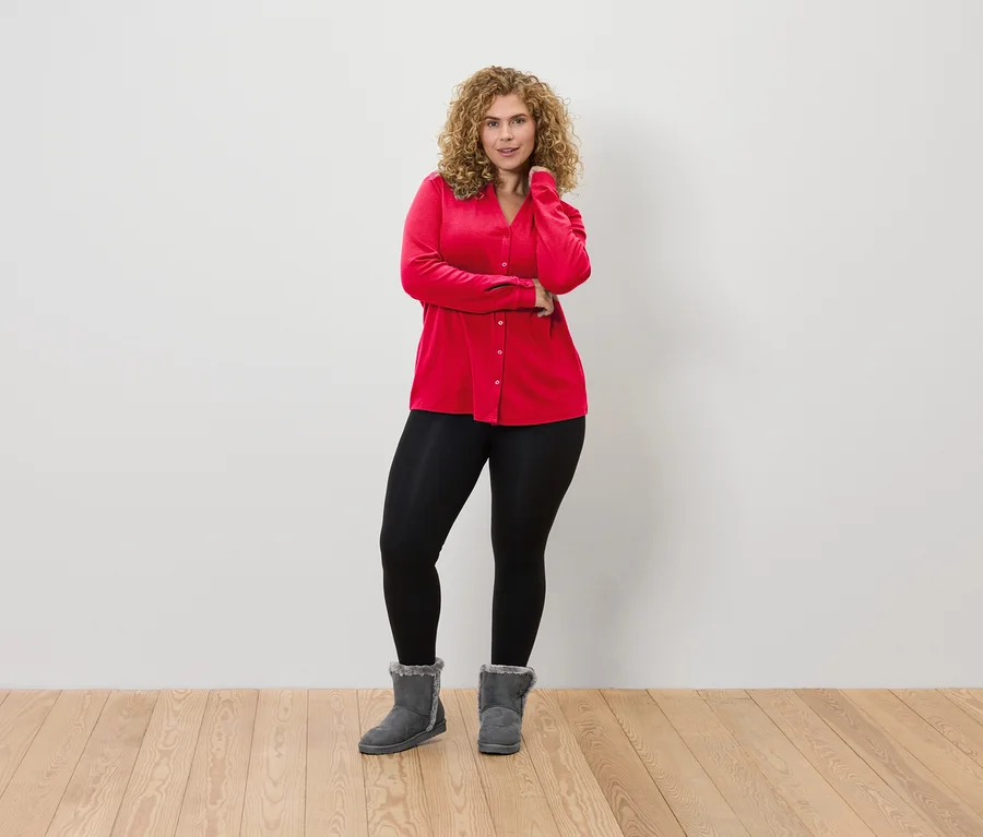Eine Frau mit lockigem Haar posiert in einer roten Bluse, schwarzen Leggings und grauen Stiefeln vor einer weißen Wand.