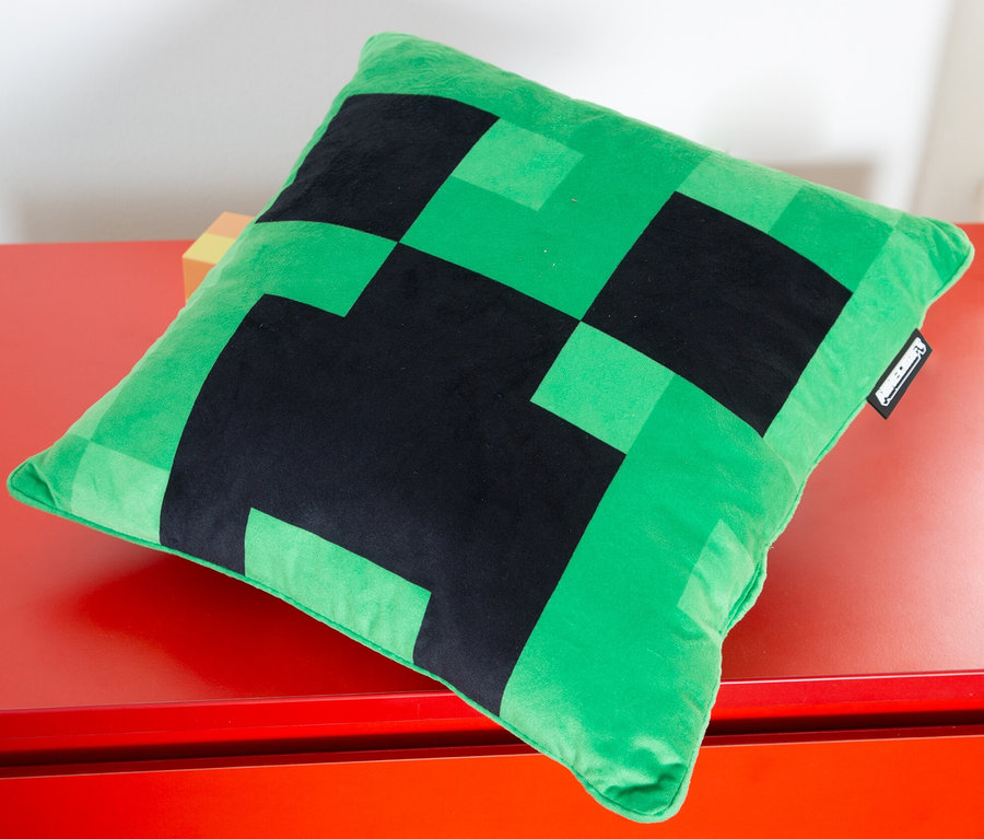 Ein grünes MINECRAFT Dekokissen, Creeper, liegt auf einer roten Oberfläche.