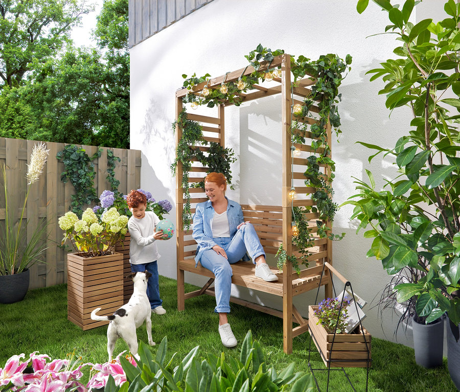 Eine Frau sitzt auf einer Gartenbank mit Rosenbogen, während ein Kind mit einem Ball spielt. Eine Solar-Lichterkette »Glühbirnen« hängt über der Bank. Im Garten stehen 2 Pflanzsäulen »Tinus« und ein Kräuterkasten mit Tragegestell »Elin«.
