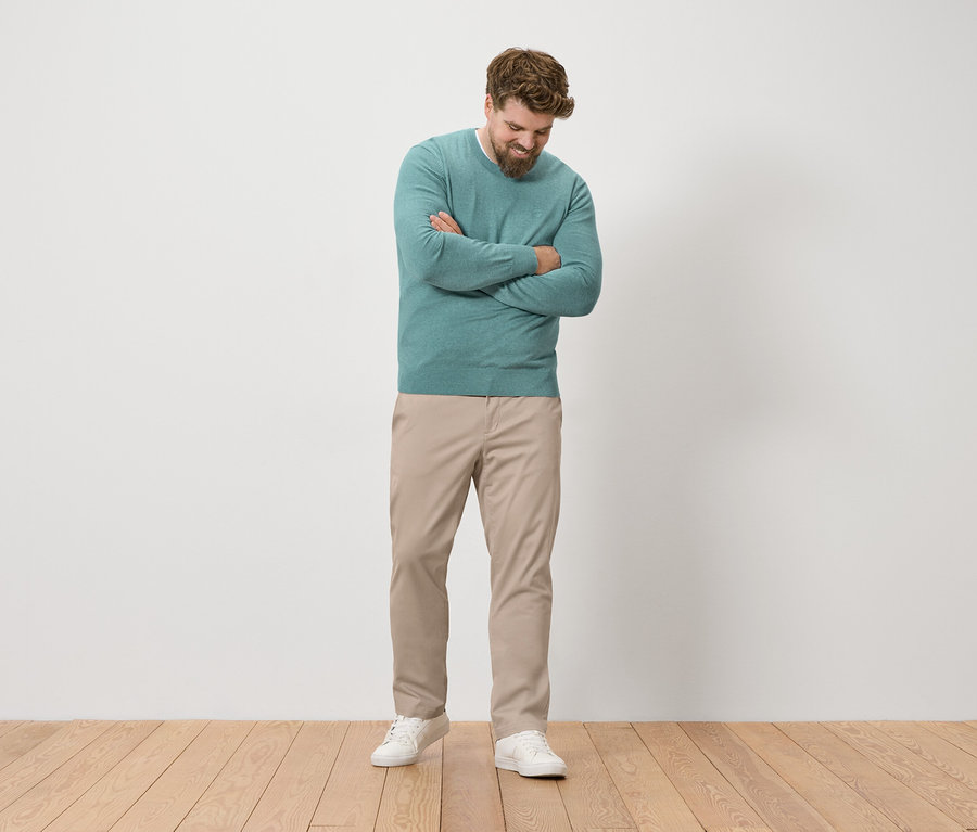 Mann mit Bart steht mit verschränkten Armen und trägt einen grünen Pullover, beige Hosen und weiße Turnschuhe.