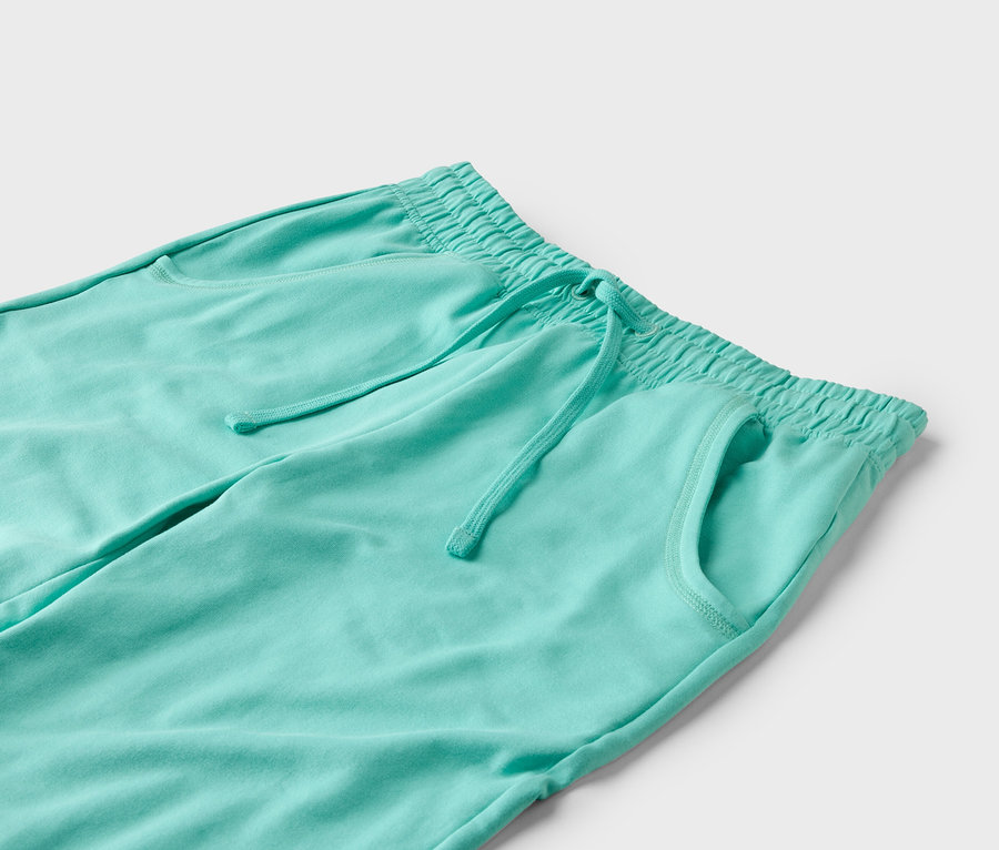 Nahaufnahme einer türkisfarbenen Jogginghose mit Kordelzug und Taschen vor einem weißen Hintergrund.