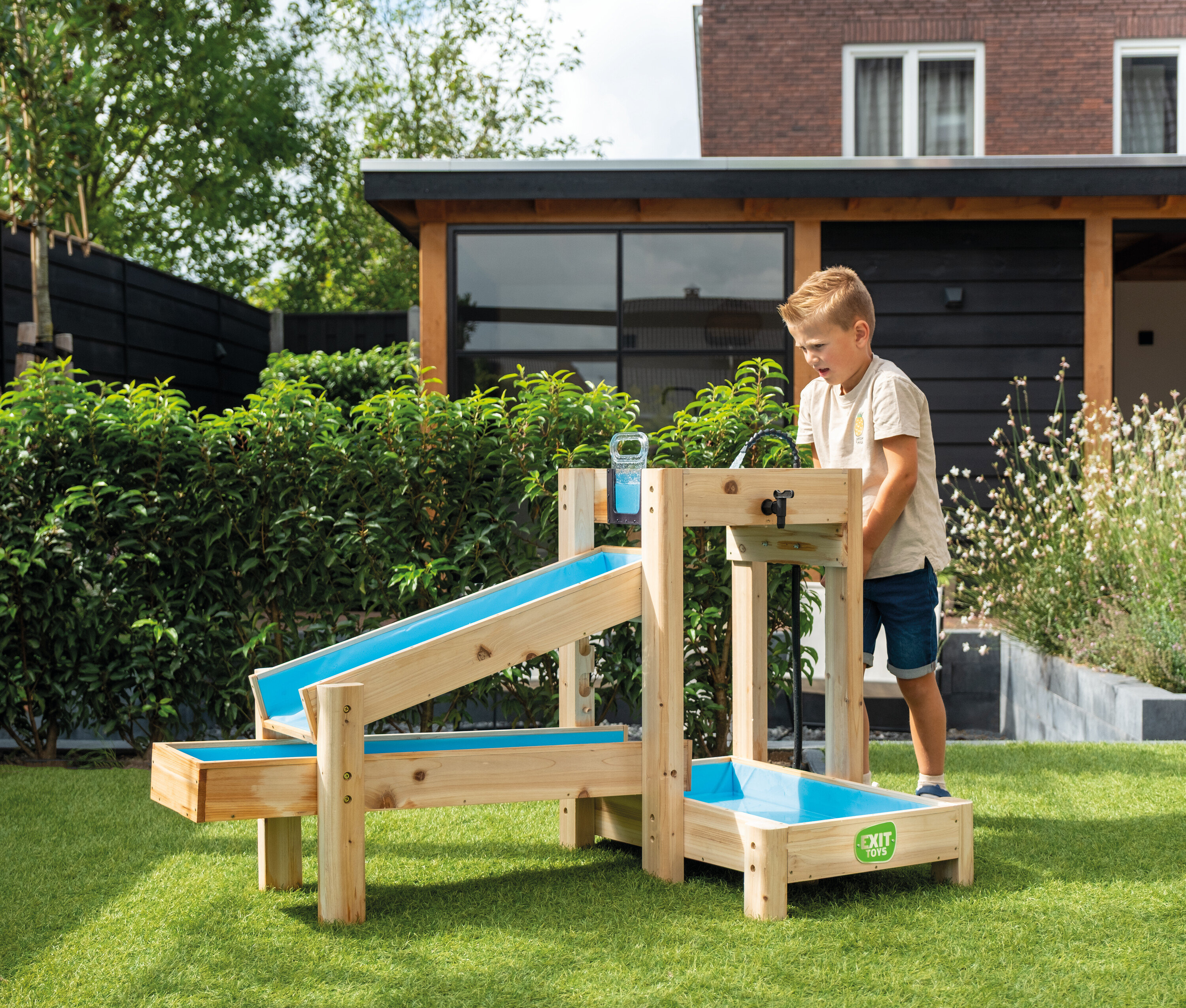 Junge spielt mit einer hölzernen Wasserspielbahn im Garten. Ein Glas Wasser steht bereit.