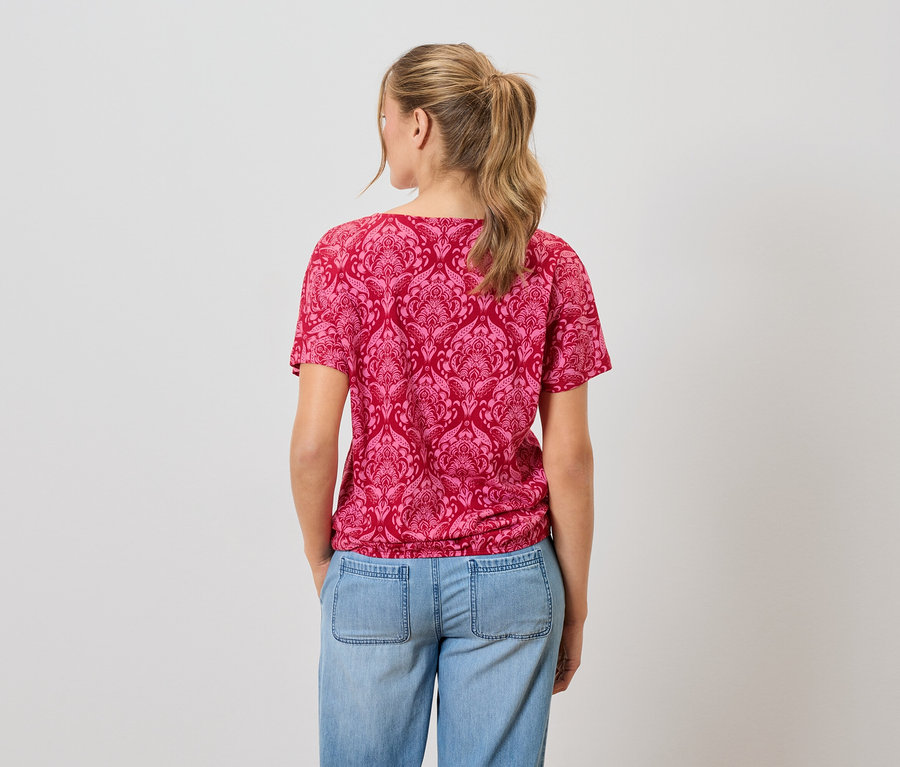 Eine Frau mit blonden Haaren trägt ein rot gemustertes T-Shirt und blaue Jeans.