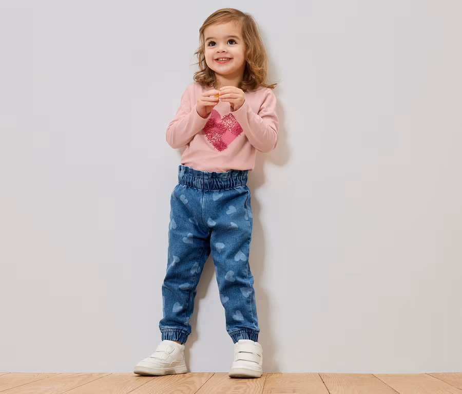 Ein kleines Mädchen mit blonden Haaren steht vor einer Wand und trägt ein rosa Langarmshirt mit einem Herz und blaue Jeans mit Herzen.