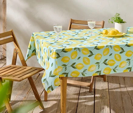 Ein Tisch mit einer hellblauen Zitronen-Tischdecke, zwei Gläsern, Zitronen auf einem Teller und einer Topfpflanze.