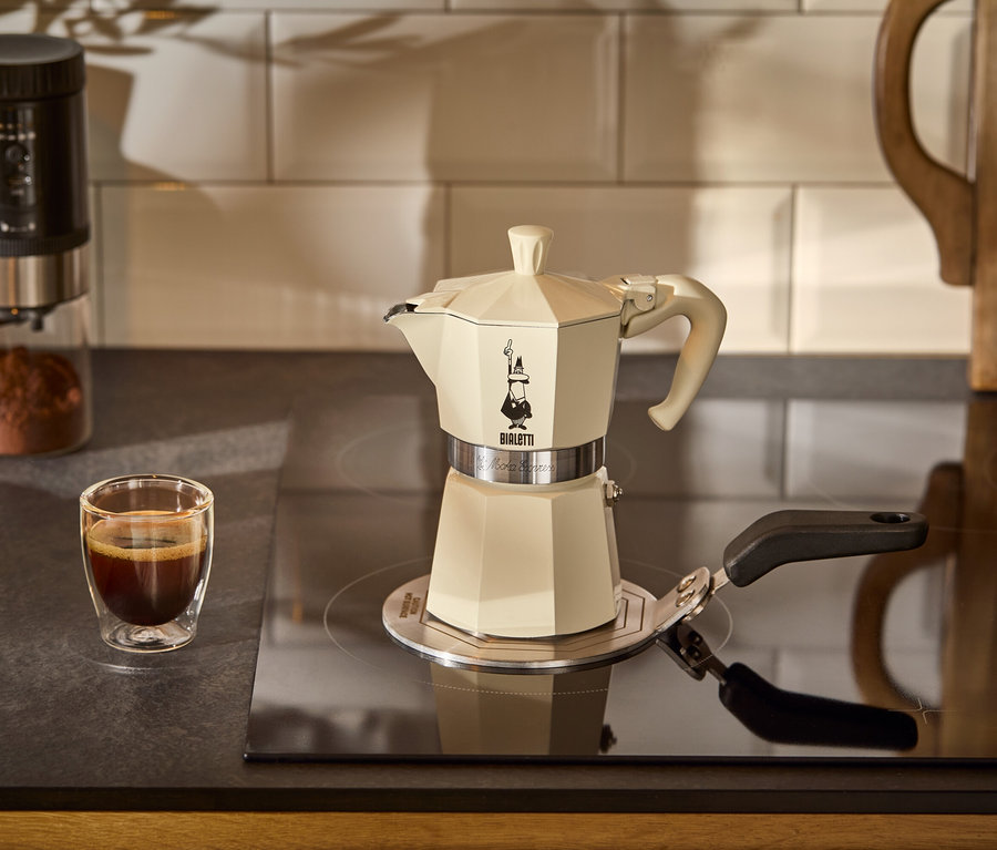 Eine Bialetti Moka Express, 190 ml, Crema steht auf einer Bialetti Adapterplatte Induktion auf einem Kochfeld. Im Hintergrund befindet sich eine Tasse Kaffee.
