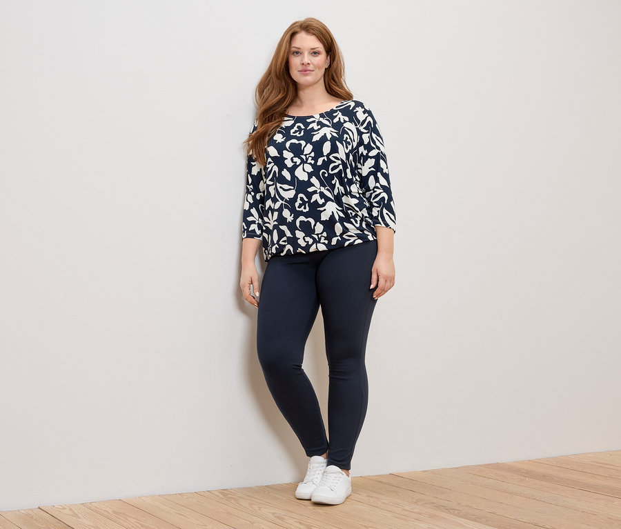 Eine Frau mit langen roten Haaren steht in einem marineblau-weißen Oberteil mit Blumenmuster, marineblauen Leggings und weißen Turnschuhen.