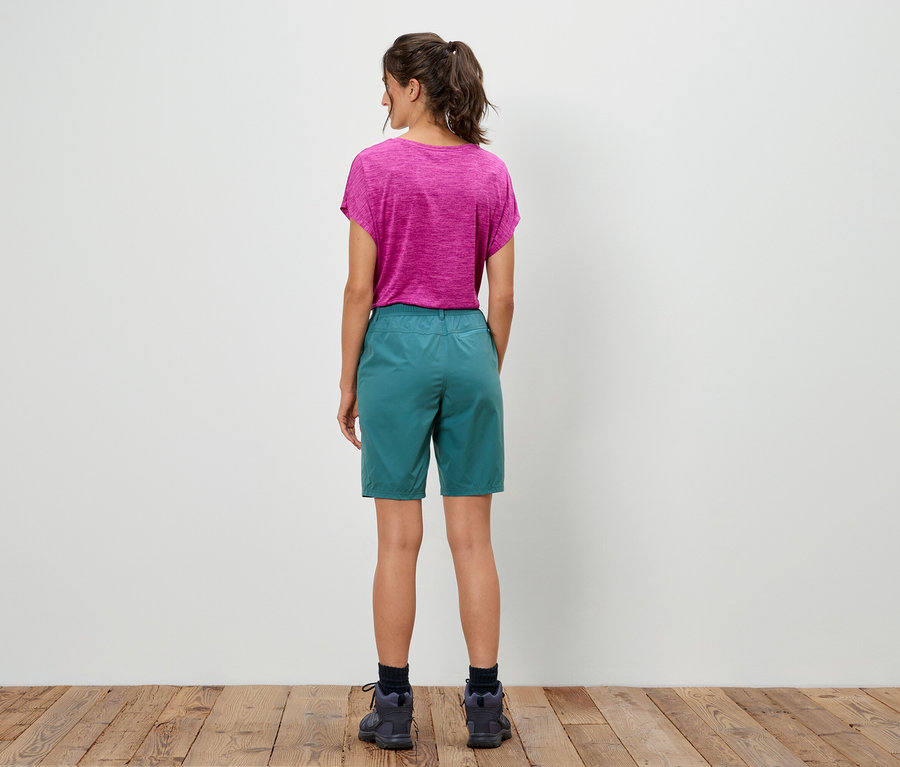 Eine Frau steht mit dem Rücken zur Kamera, sie trägt ein pinkes T-Shirt und eine grüne Shorts.