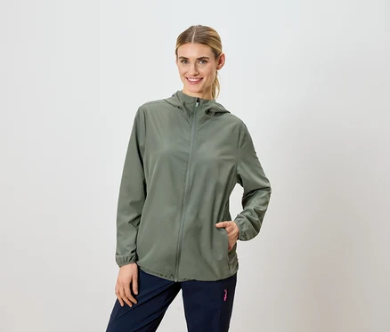 Frau mit blonden Haaren trägt eine olivgrüne Kapuzenjacke und marineblaue Hosen.
