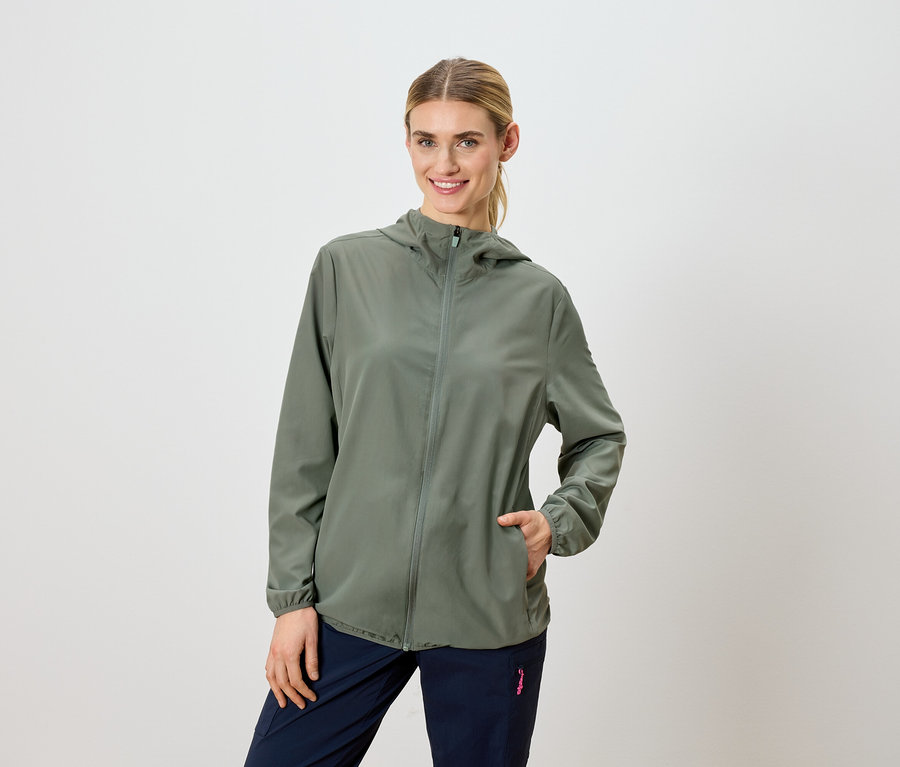 Frau mit blonden Haaren trägt eine olivgrüne Kapuzenjacke und marineblaue Hosen.