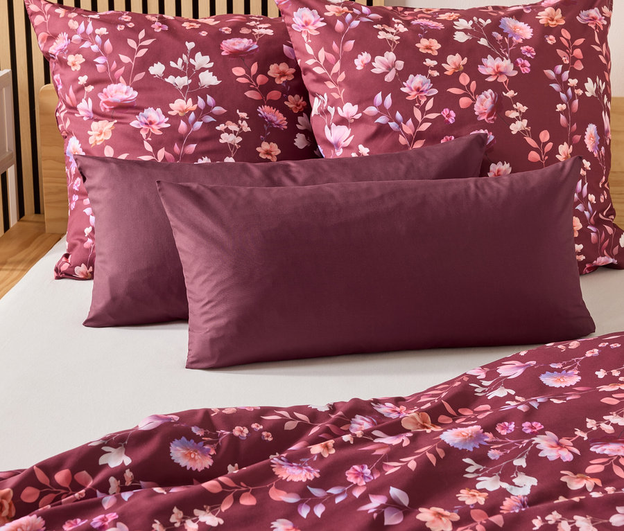 Auf einem Bett liegen zwei burgunderfarbene Kissen und zwei Kopfkissen mit Blumenmuster. Eine burgunderfarbene Bettdecke liegt am Fußende.