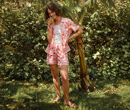 Frau mit Glas in CALIDA »Favorites Beach« Shorts und passendem Hemd, stehend an einem Baum.