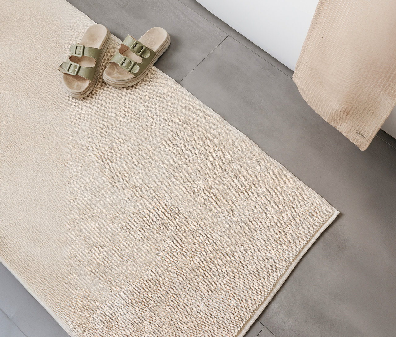 Beige benuta Badematte »Ole« (50 x 80 cm) liegt auf grauem Boden, daneben Sandalen und ein beiges Handtuch.