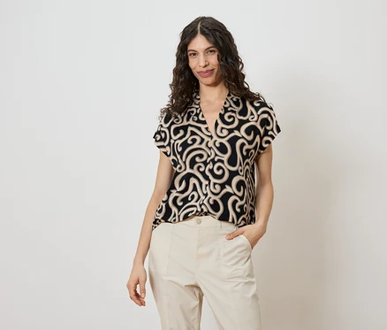 Frau mit lockigem Haar trägt eine schwarze Bluse mit beigem Muster und beige Hosen.