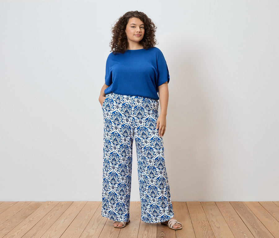 Frau mit lockigem Haar, die ein blaues Oberteil und eine blau-weiss gemusterte Hose trägt.