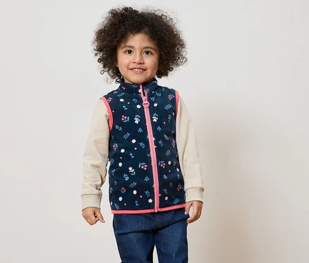 Ein lächelndes Kind mit lockigem Haar trägt eine marineblaue Weste mit Blumenmuster und rosa Reißverschluss über einem beigen Oberteil und Jeans.