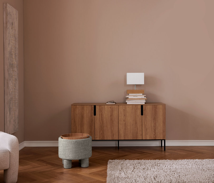 Ein Wohnzimmer mit hellbraunen Wänden, einem Fischgrätparkettboden und einem hellen, zotteligen Teppich. Ein Holzsideboard mit einer weißen rechteckigen Lampe und Büchern steht an der Wand. Links davon ein grauer runder Hocker mit Holztablett und ein heller Sessel ist links teilweise sichtbar.