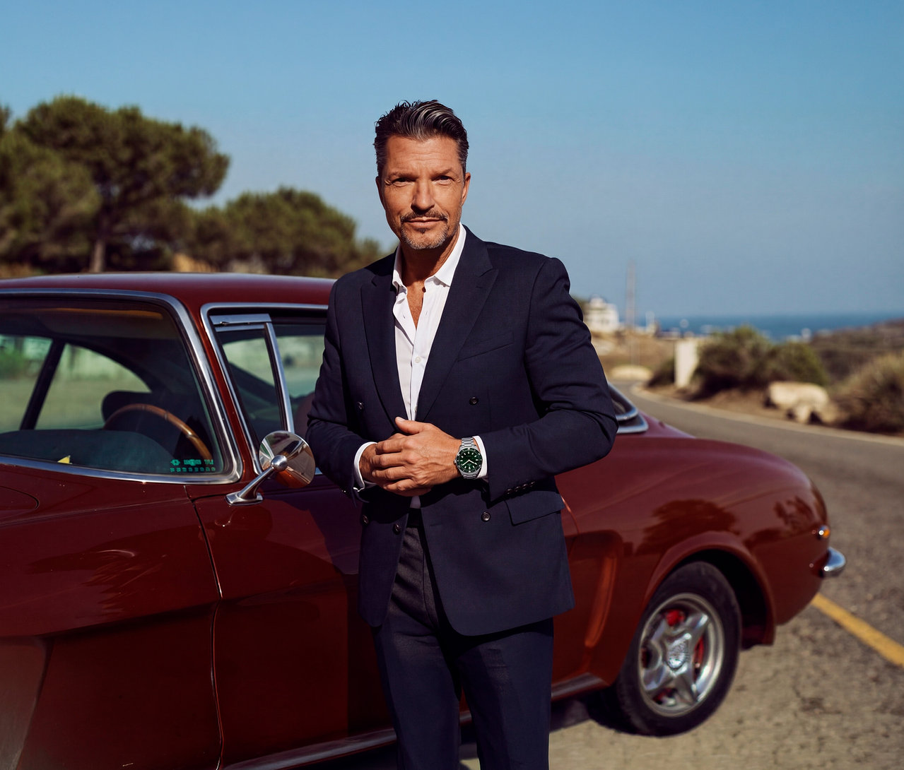 Ein Mann in einem dunkelblauen Anzug und weißem Hemd steht neben einem roten Oldtimer. Er trägt eine grüne Kienzle Uhr »Taucher Quarz« am Handgelenk, im Hintergrund ist das Meer zu sehen.