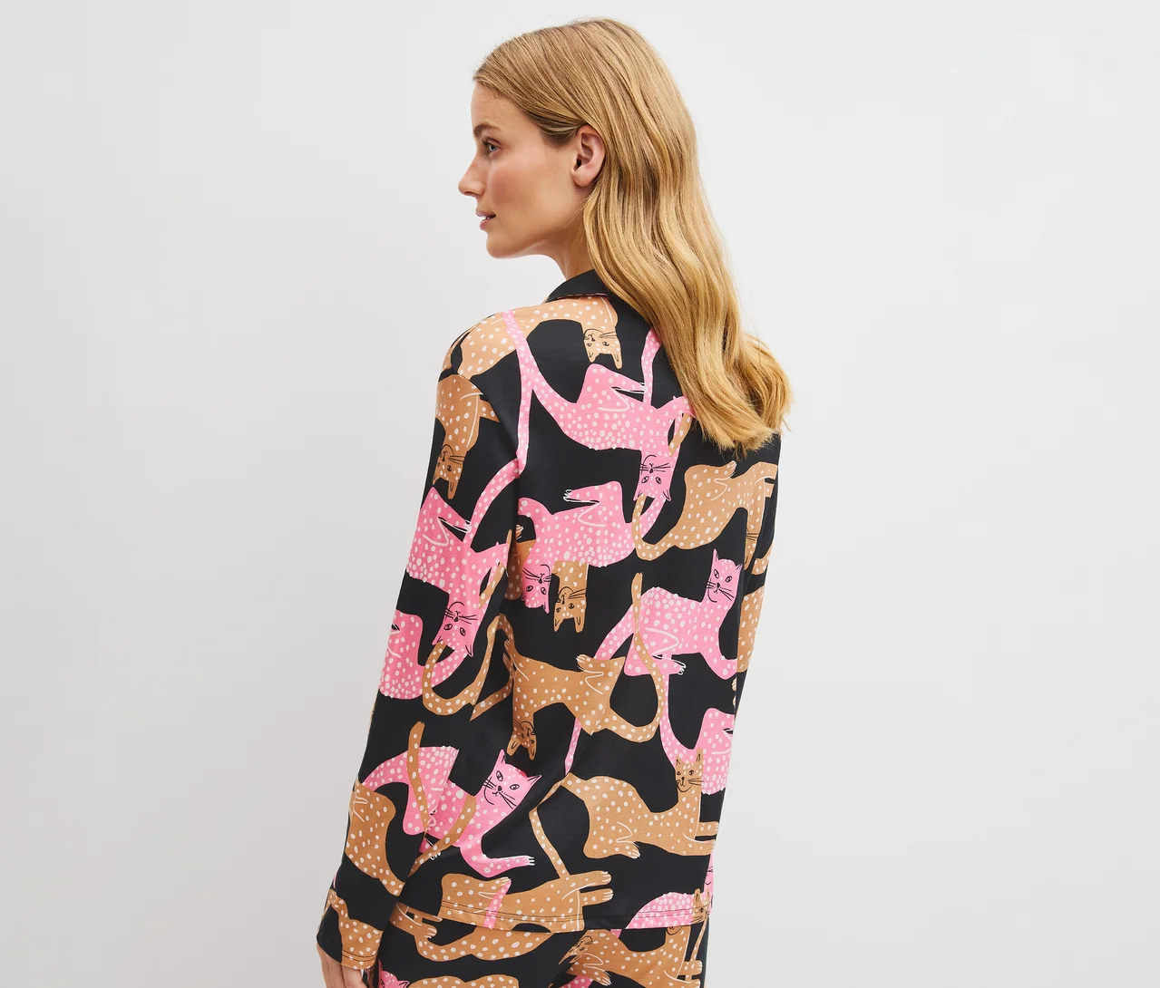 Frau von hinten gesehen, nach links blickend, trägt ein schwarzes Langarmshirt mit einem Muster aus rosa und braunen Katzen.
