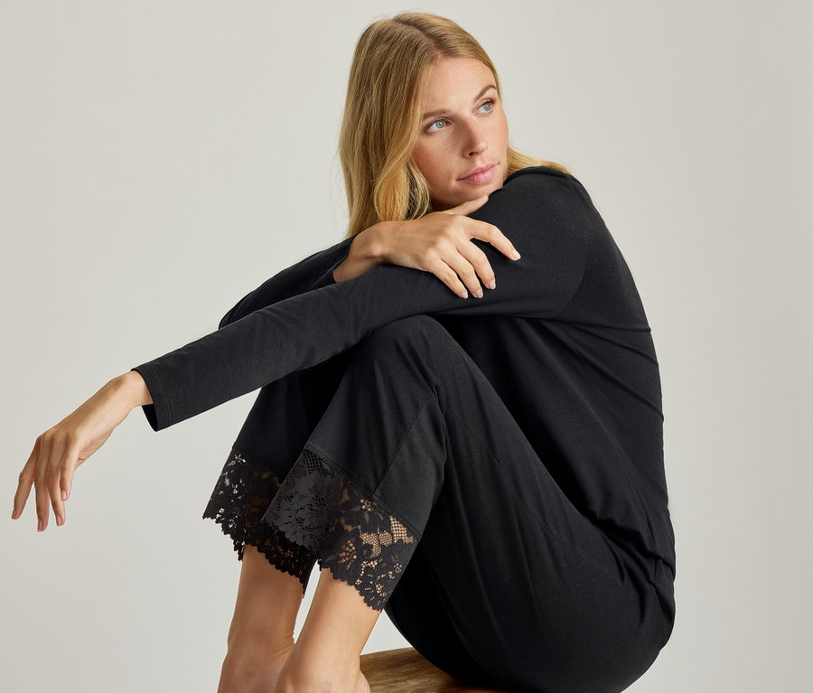 Frau sitzt in einem schwarzen Pyjama-Set mit langen Ärmeln und Spitzenbesatz an den Hosenbeinen.