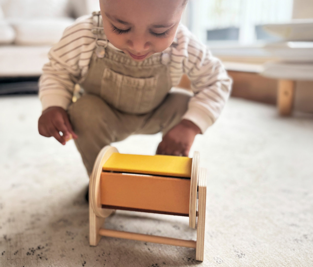 Ein kleines Kind in gestreiftem Shirt und beigem Overall hockt auf einem Teppich und spielt mit dem small foot Montessori-Bundle 3, einem Holzspielzeug mit gelben und orangen Elementen.