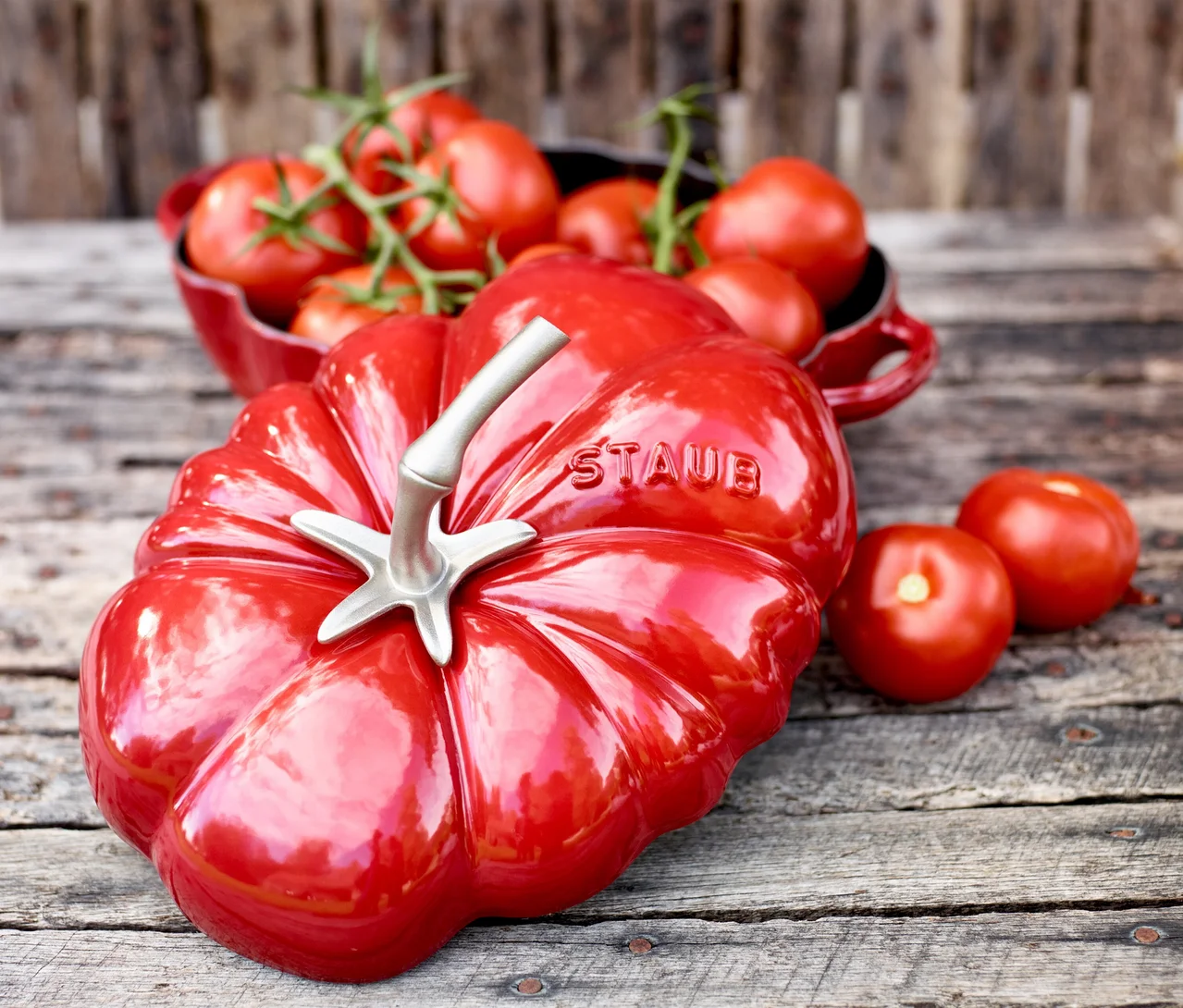 Roter, tomatenförmiger STAUB Gusseisen-Bräter »Tomate« auf Holz, umgeben von frischen Tomaten in einer Schale und lose.