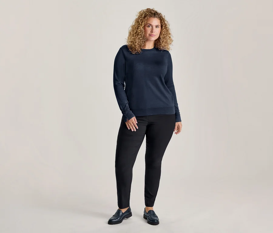 Frau mit lockigem blondem Haar trägt einen dunkelblauen Feinstrickpullover, eine schwarze Bengalin-Hose und schwarze Loafer.