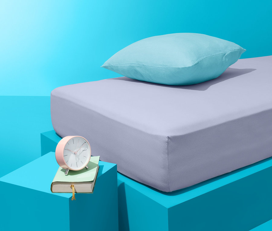 Auf blauem Hintergrund befinden sich ein Bett mit lila Bettlaken und ein blaues Kissen. Neben dem Bett steht ein blauer Tisch mit einem Buch und einem rosa Wecker.