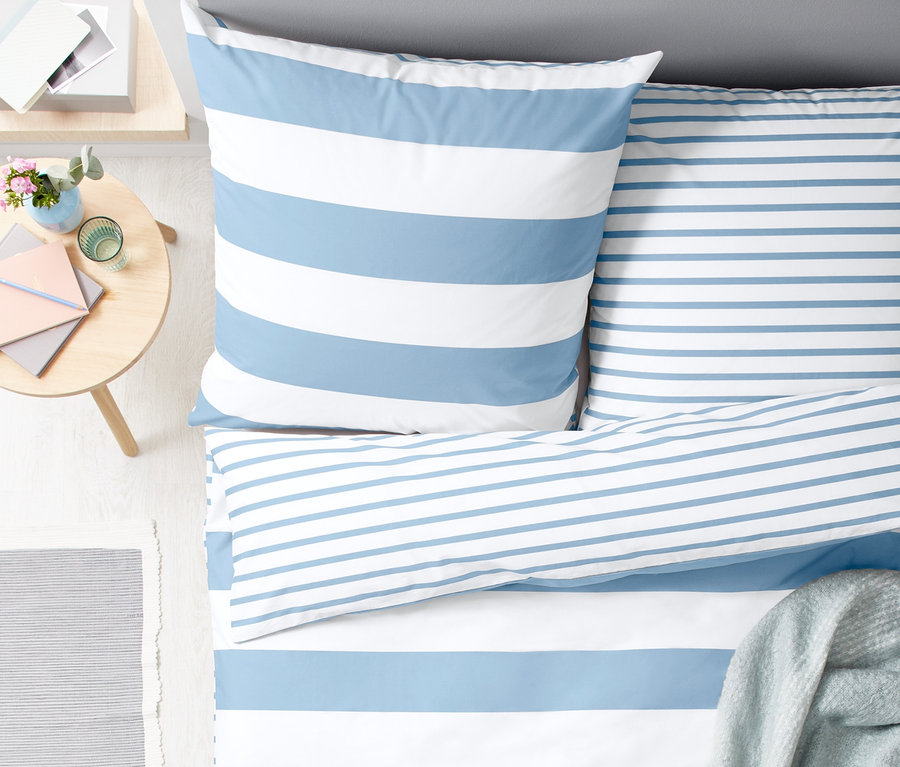Draufsicht auf ein Doppelbett mit Perkal-Wendebettwäsche mit blauen und weißen Streifen. Eine graue Decke liegt auf dem Bett.
