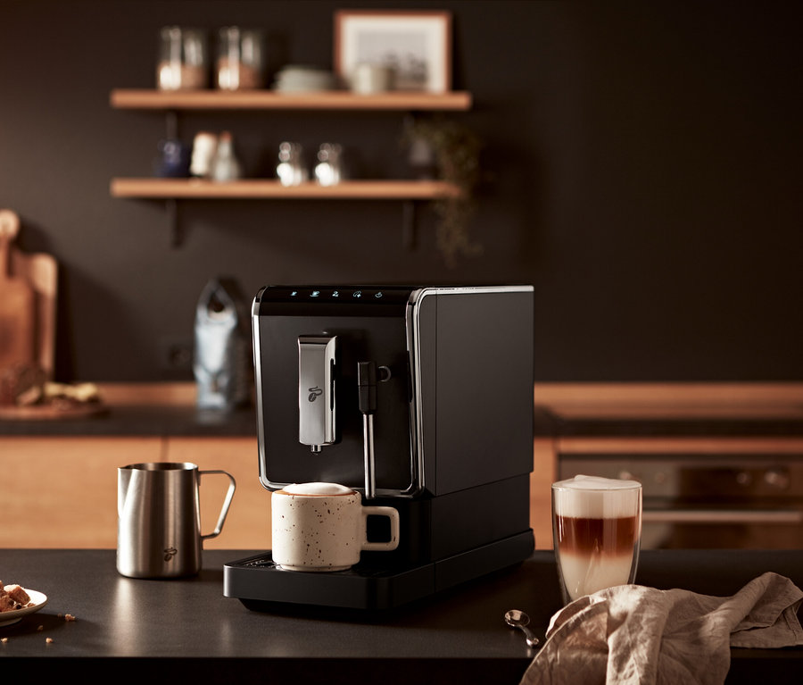 Auf einem Tisch steht ein »Esperto Latte« Tchibo Kaffeevollautomat, Anthrazit, neben einer Tasse Kaffee und einem Glas Latte.