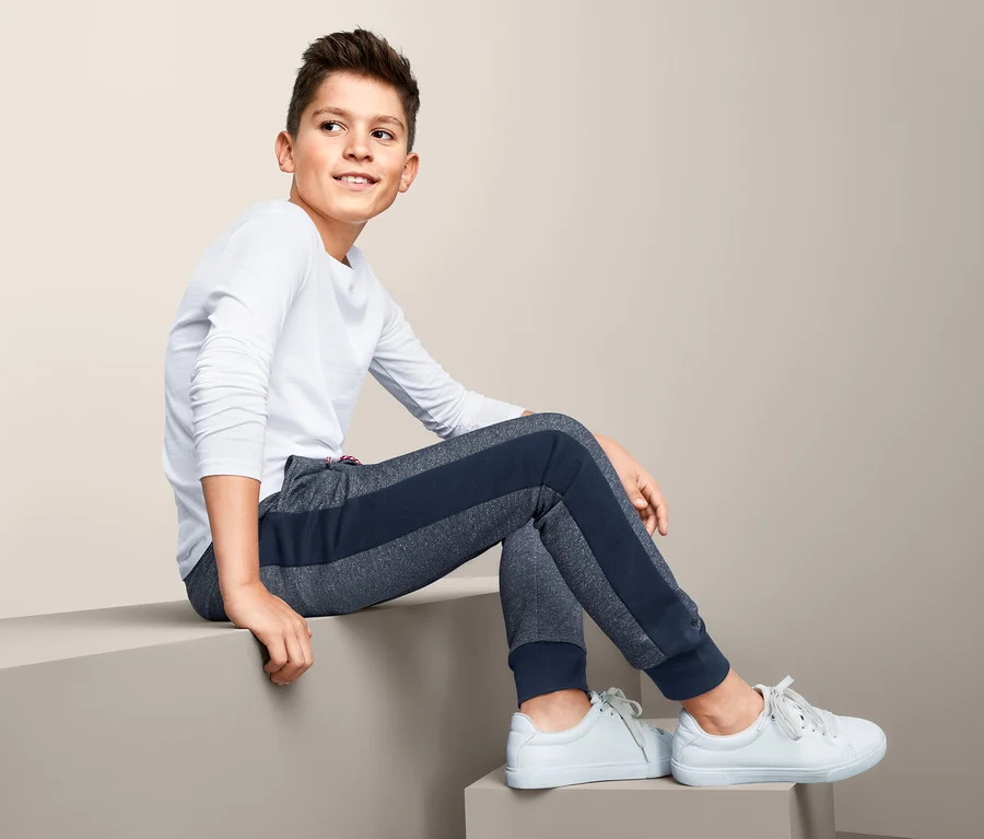 Ein Junge sitzt auf einer Kante und trägt eine Sweathose und ein weißes Langarmshirt.