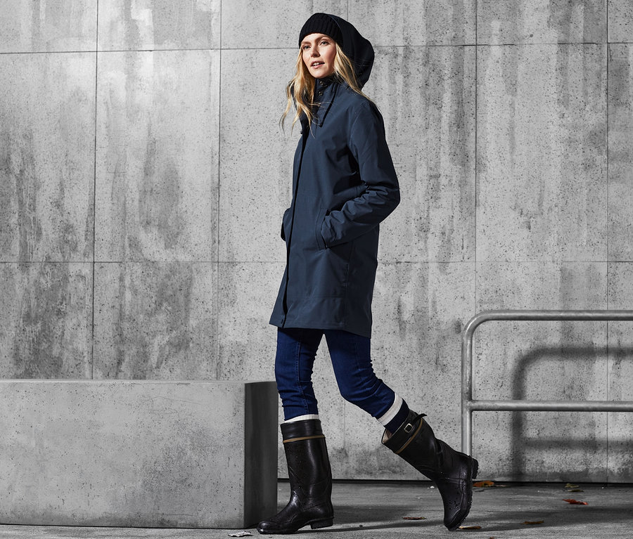 Frau geht in Hardshell-Regenparka, Jeans und Gummistiefeln.