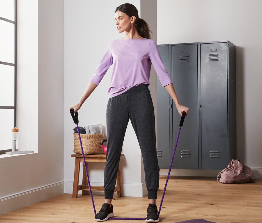 Frau trainiert mit einem Expander auf einer Matte auf dem Boden. Sie trägt eine Trainingshose.