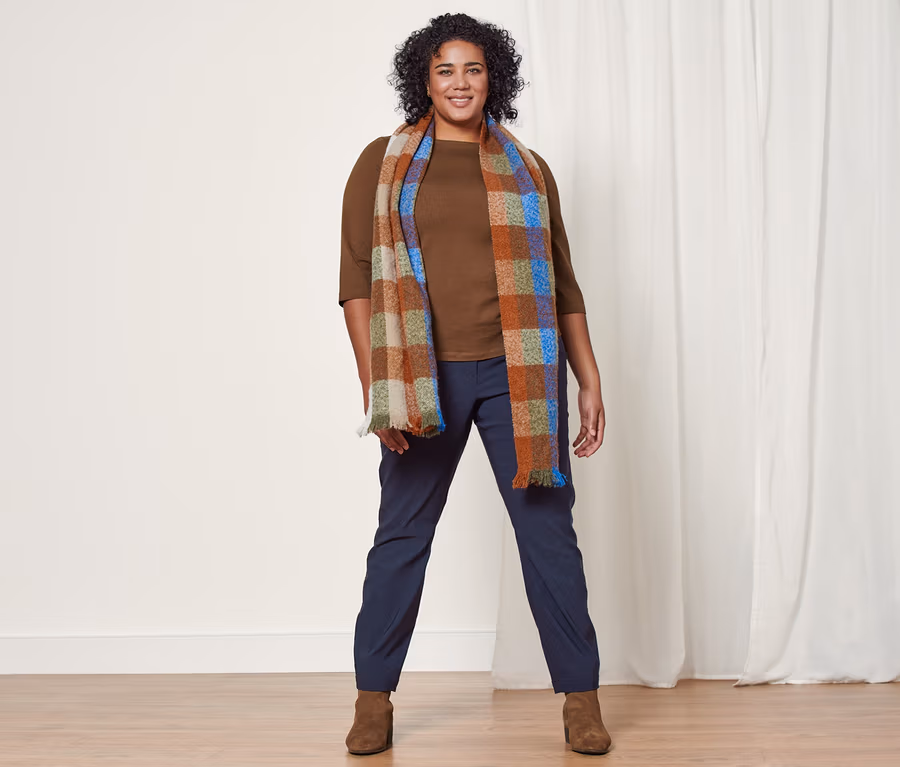 Eine Frau steht und trägt ein rostbraunes Shirt mit 3/4-Arm, einen karierten Webschal, eine dunkelblaue, konfektionierte Bengalinhose und Stiefeletten aus Veloursleder.