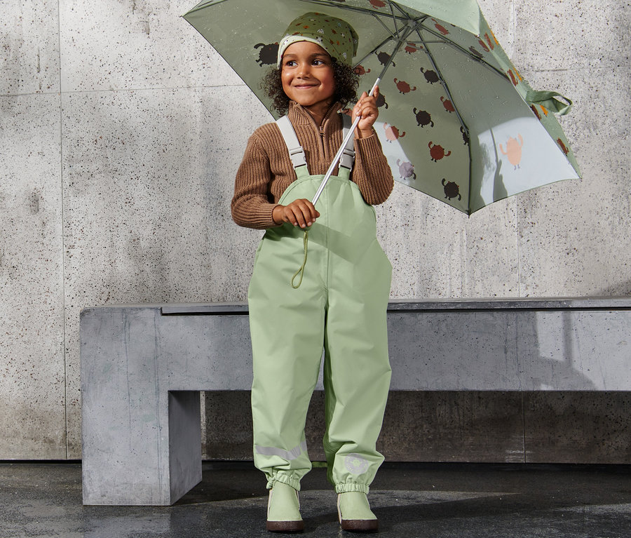 Ein lächelndes Mädchen hält einen bedruckten Regenschirm und trägt eine Regenhose.