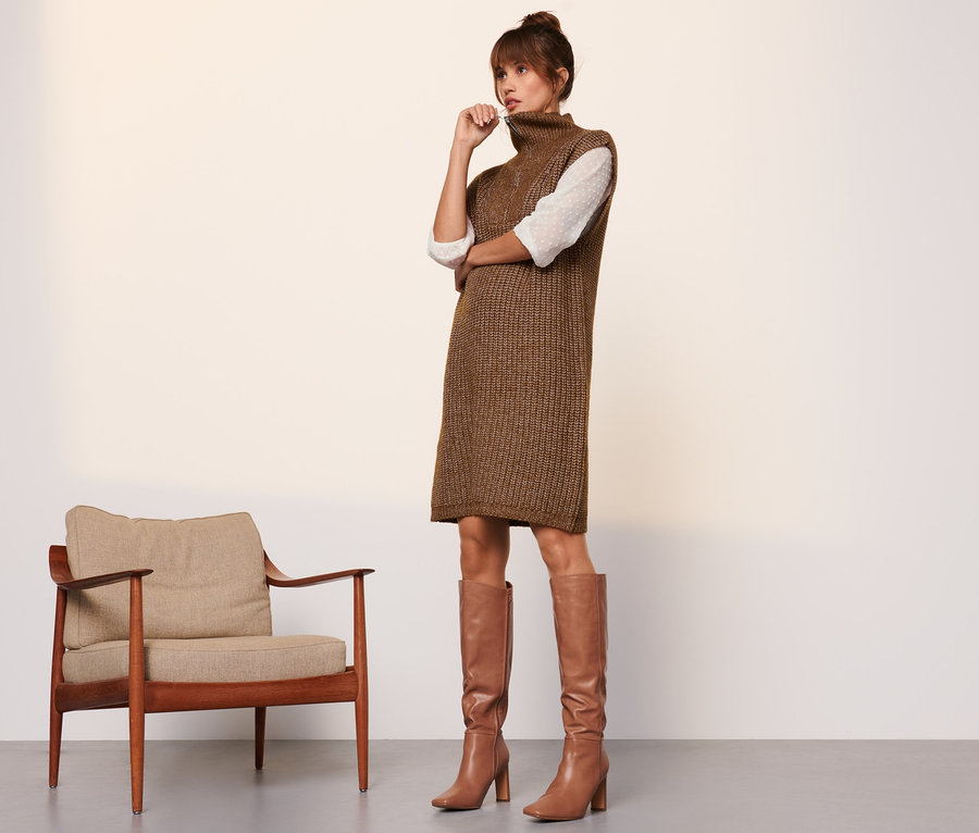Frau steht neben Sessel, trägt braunes Strickkleid und Stiefel.