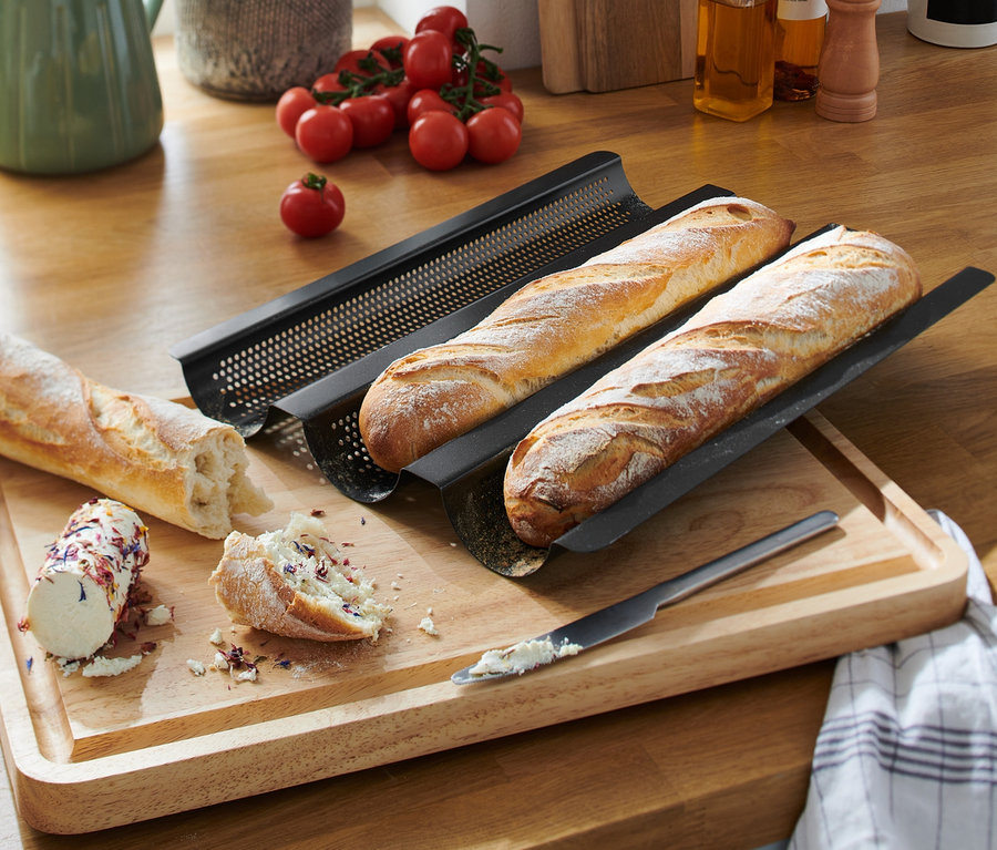 Zwei Baguettes liegen in einem schwarzen, gelochten Baguette-Backblech auf einem Holzbrett. Daneben liegt ein aufgeschnittenes Baguette mit Aufstrich und ein Messer.