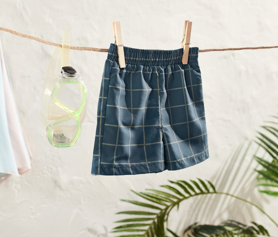 Kleinkind-Boardshorts hängen an einer Wäscheleine, daneben eine Taucherbrille und ein Strandschirm.
