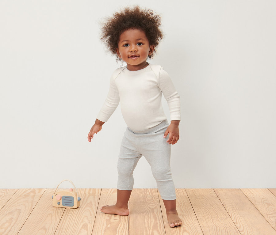 Ein Kind steht auf einem Holzboden und trägt 3 Baby-Ripp-Langarmbodys und graue Leggings.