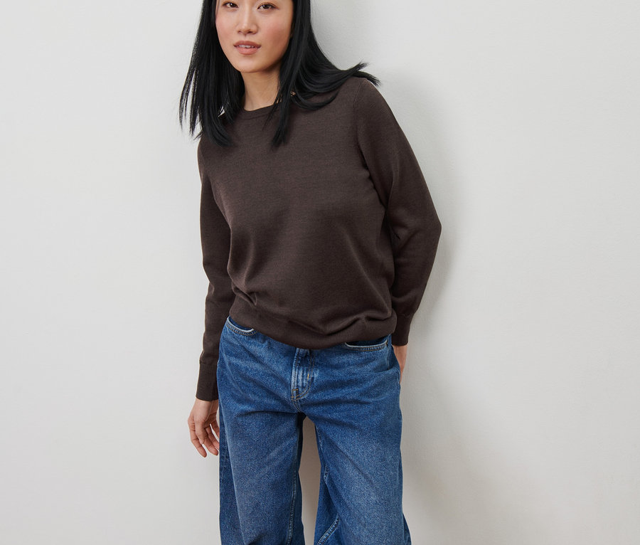 Asiatische Frau lehnt an einer weißen Wand und trägt einen braunen Merino-Feinstrickpullover und blaue Jeans.