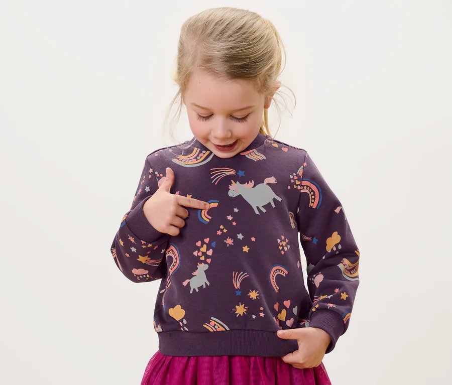 Mädchen zeigt auf Kinder-Sweater mit Alloverprint von Einhörnern, Sternen und Regenbögen.