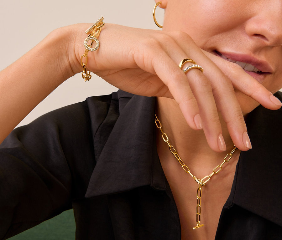 Frau trägt goldenen Ring Doppel-Welle mit Kristallen, Kette und Armband Toggle mit Kristallbesatz und goldfarbene Creolen.