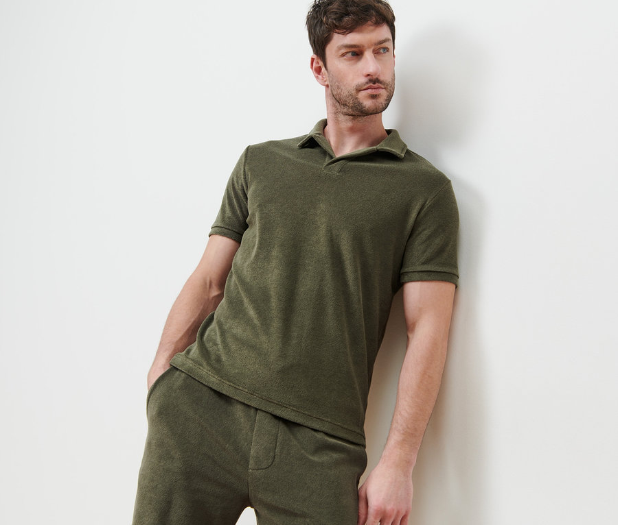 Mann lehnt an Wand, trägt Frottee-Poloshirt, khaki.